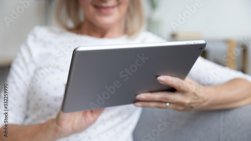 Close up pleasant elder female retiree holding digital tablet, sitting on couch at home. Positive middle aged woman searching information or shopping online. Older generation easy technology usage.