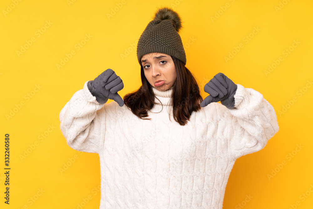 Young woman with winter hat over isolated yellow background showing thumb down Young woman with winter hat over isolated yellow background showing thumb down