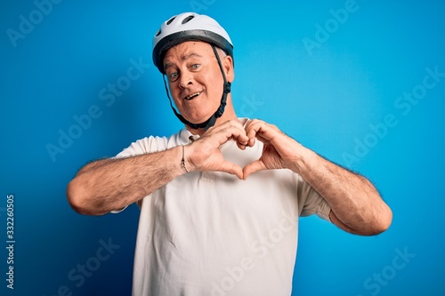Middle age hoary cyclist man wearing bike security helmet over isolated blue background smiling in love showing heart symbol and shape with hands. Romantic concept.
