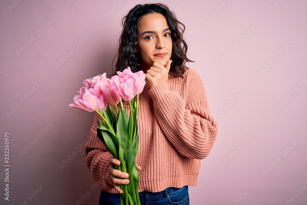 Young beautiful romantic woman with curly hair holding bouquet of pink tulips looking confident at the camera smiling with crossed arms and hand raised on chin. Thinking positive. Young beautiful romantic woman with curly hair holding bouquet of pink tulips looking confident at the camera smiling with crossed arms and hand raised on chin. Thinking positive.
