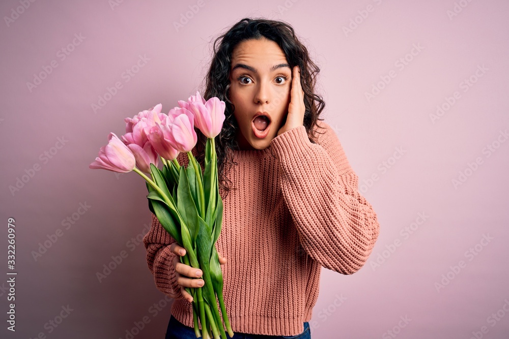Young beautiful romantic woman with curly hair holding bouquet of pink tulips afraid and shocked, surprise and amazed expression with hands on face Young beautiful romantic woman with curly hair holding bouquet of pink tulips afraid and shocked, surprise and amazed expression with hands on face