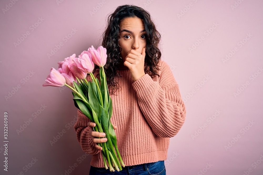 Young beautiful romantic woman with curly hair holding bouquet of pink tulips shocked covering mouth with hands for mistake. Secret concept. Young beautiful romantic woman with curly hair holding bouquet of pink tulips shocked covering mouth with hands for mistake. Secret concept.