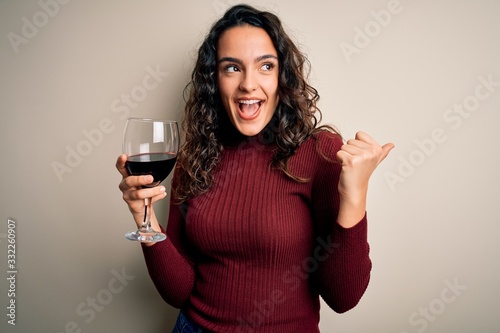 Young beautiful woman with curly hair drinking glass of red wine over white background pointing and showing with thumb up to the side with happy face smiling photo