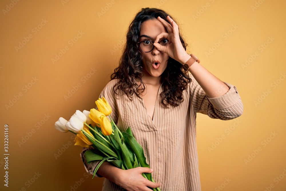 Young beautiful romantic woman with curly hair holding bouquet of yellow tulips doing ok gesture shocked with surprised face, eye looking through fingers. Unbelieving expression. Young beautiful romantic woman with curly hair holding bouquet of yellow tulips doing ok gesture shocked with surprised face, eye looking through fingers. Unbelieving expression.