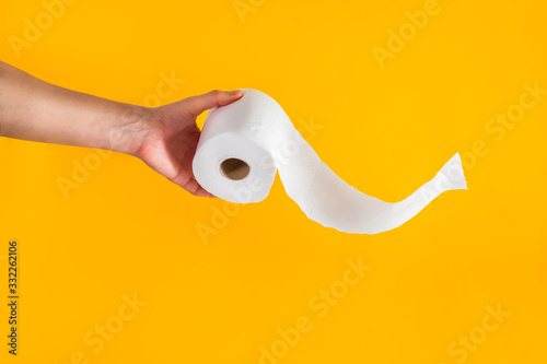 Close up caucasian male hand holding or giving a roll of toilet paper with flying tissue on bright yellow background. Crisis product lack concept. Copy space. photo