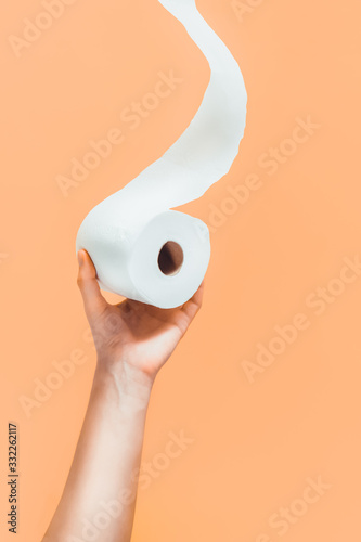 Close up caucasian male hand pulling or holding a roll of toilet paper from buttom on light orange, beige background. Crisis product lack concept. Modern monochrome vertical card. Copy space.