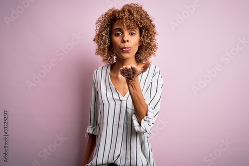 Beautiful african american woman with curly hair wearing striped t-shirt over pink background looking at the camera blowing a kiss with hand on air being lovely and sexy. Love expression.