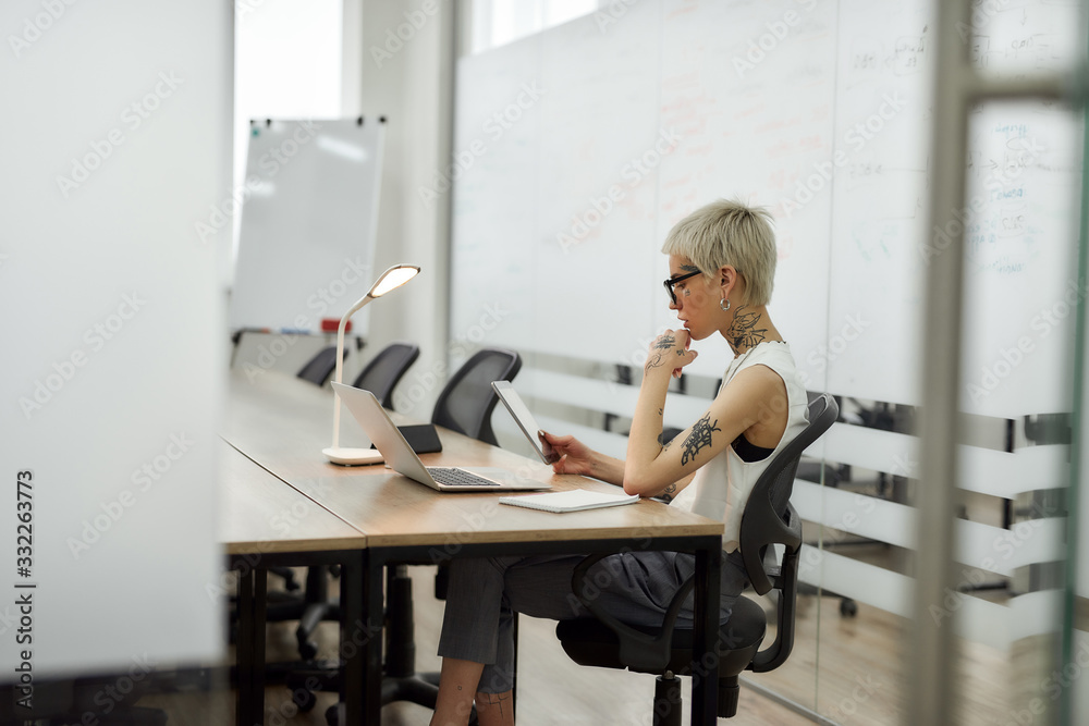 Working alone. Young tattooed business lady in classic wear with short haircut using digital tablet while sitting in the conference room in the modern office