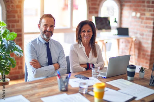 Middle age beautiful business workers working together using laptop at the office happy face smiling with crossed arms looking at the camera. Positive person.