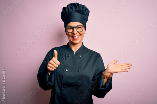 Middle age brunette chef woman wearing cooker uniform and hat over isolated pink background Showing palm hand and doing ok gesture with thumbs up, smiling happy and cheerful photo