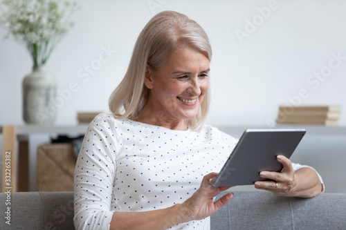 Head shot happy older lady relaxing on comfortable sofa, holding computer tablet in hands, watching movie. Smiling middle aged woman reading book online or making purchases in internet store at home.
