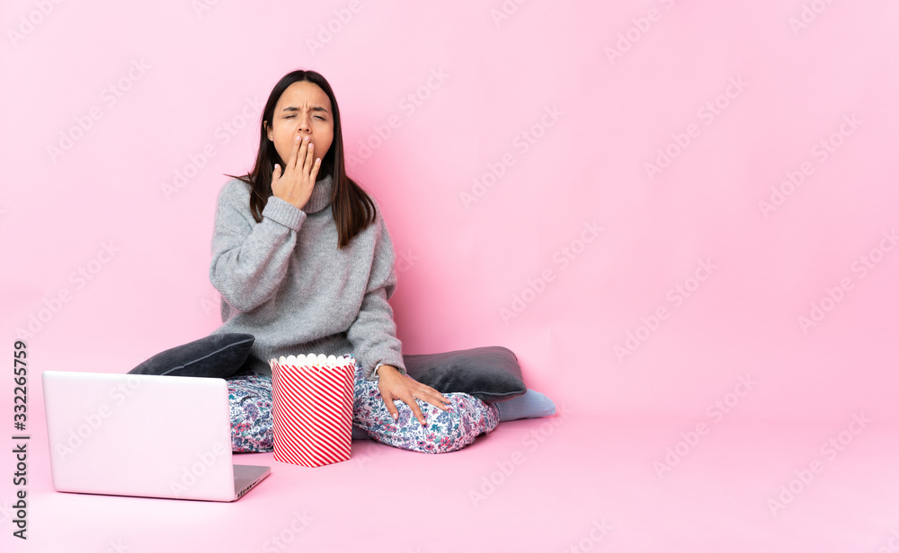 Young mixed race woman eating popcorn while watching a movie on the laptop yawning and covering wide open mouth with hand Young mixed race woman eating popcorn while watching a movie on the laptop yawning and covering wide open mouth with hand