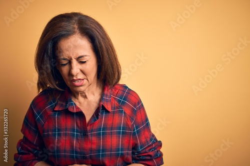 Middle age beautiful woman wearing casual shirt standing over isolated yellow background with hand on stomach because indigestion, painful illness feeling unwell. Ache concept.