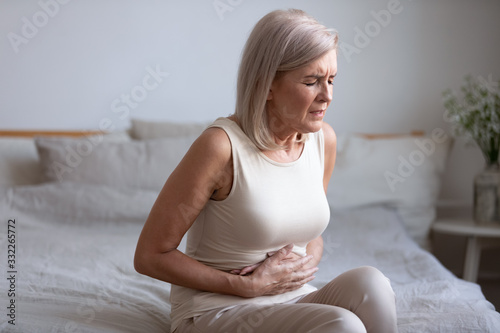 Canvas Print Unhealthy middle aged frowning woman sitting on bed, putting hands on belly, suffering from strong stomach ache