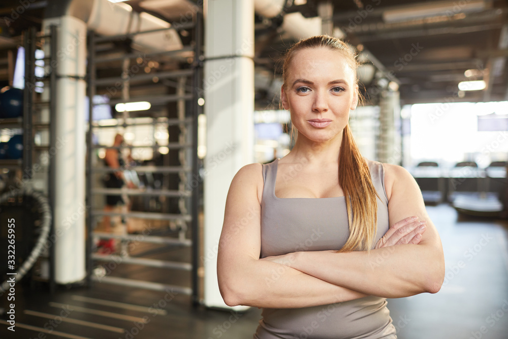 Waist up portrait of muscular woman smiling at camera while standing with arms crossed in modern gym, copy space
