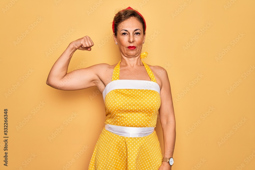 Middle age senior pin up woman wearing 50s style retro dress over yellow background Strong person showing arm muscle, confident and proud of power Middle age senior pin up woman wearing 50s style retro dress over yellow background Strong person showing arm muscle, confident and proud of power