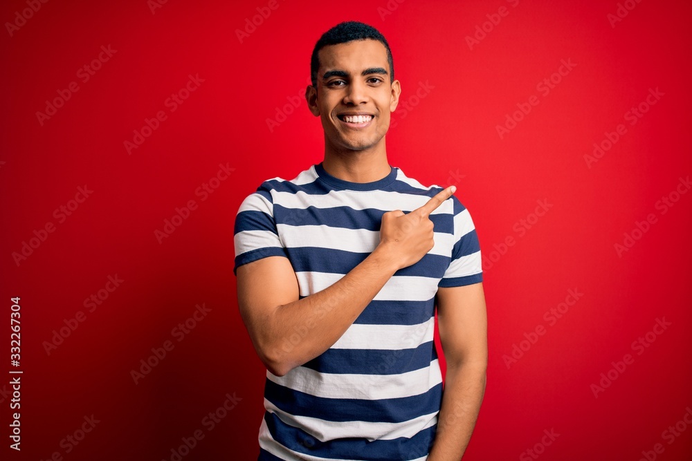 Handsome african american man wearing casual striped t-shirt standing over red background cheerful with a smile on face pointing with hand and finger up to the side with happy and natural expression Handsome african american man wearing casual striped t-shirt standing over red background cheerful with a smile on face pointing with hand and finger up to the side with happy and natural expression