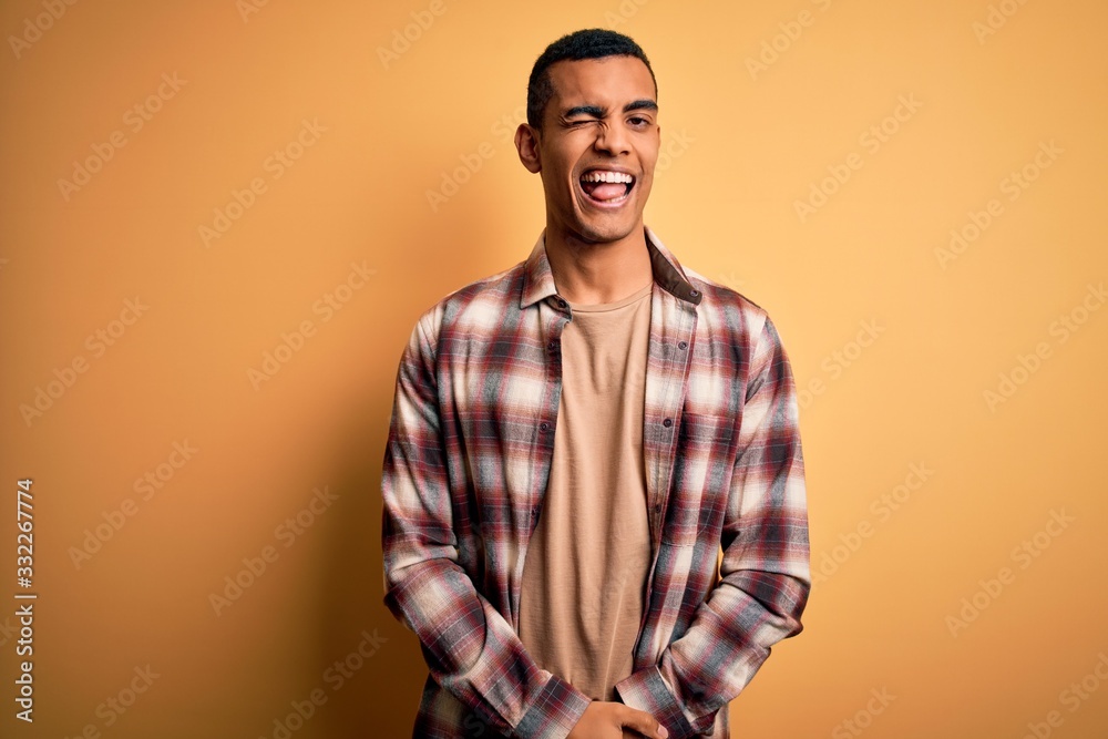 Young handsome african american man wearing casual shirt standing over yellow background winking looking at the camera with sexy expression, cheerful and happy face. Young handsome african american man wearing casual shirt standing over yellow background winking looking at the camera with sexy expression, cheerful and happy face.