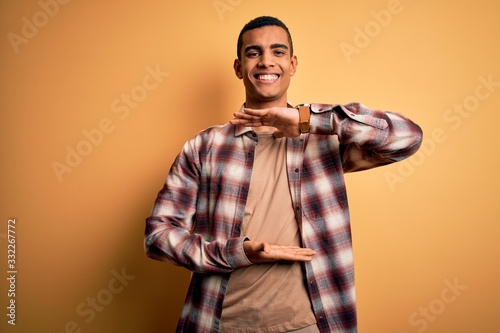 Young handsome african american man wearing casual shirt standing over yellow background gesturing with hands showing big and large size sign  measure symbol. Smiling looking at the camera. Measuring