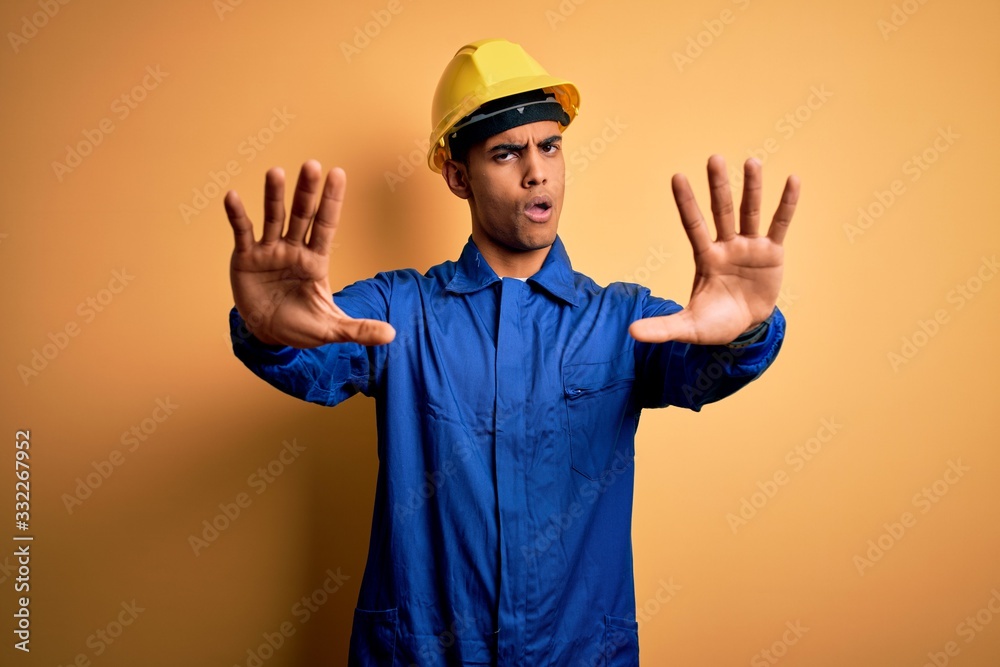 Young handsome african american worker man wearing blue uniform and security helmet doing stop gesture with hands palms, angry and frustration expression