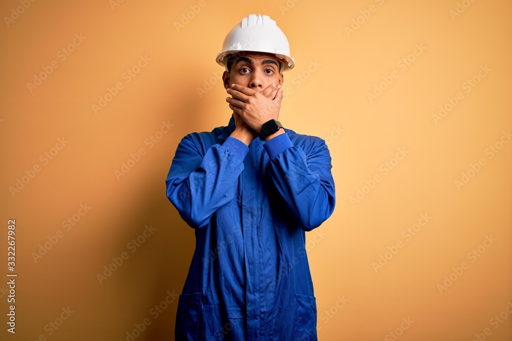 Young handsome african american worker man wearing blue uniform and security helmet shocked covering mouth with hands for mistake. Secret concept.