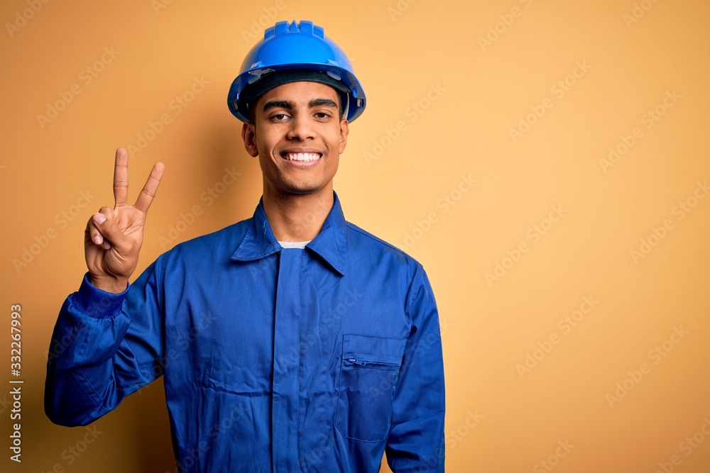 Young handsome african american worker man wearing blue uniform and security helmet smiling with happy face winking at the camera doing victory sign. Number two.