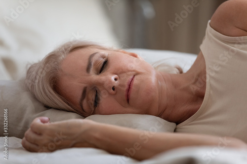 Side view serene older woman sleeping on comfortable pillow in bedroom. Head shot close up peaceful middle aged female retiree enjoying sweet dreams at home, resting alone at night or weekend morning.