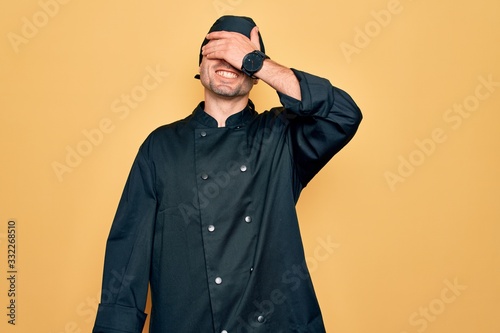 Young handsome cooker man with blue eyes wearing uniform and hat over yellow background smiling and laughing with hand on face covering eyes for surprise. Blind concept.