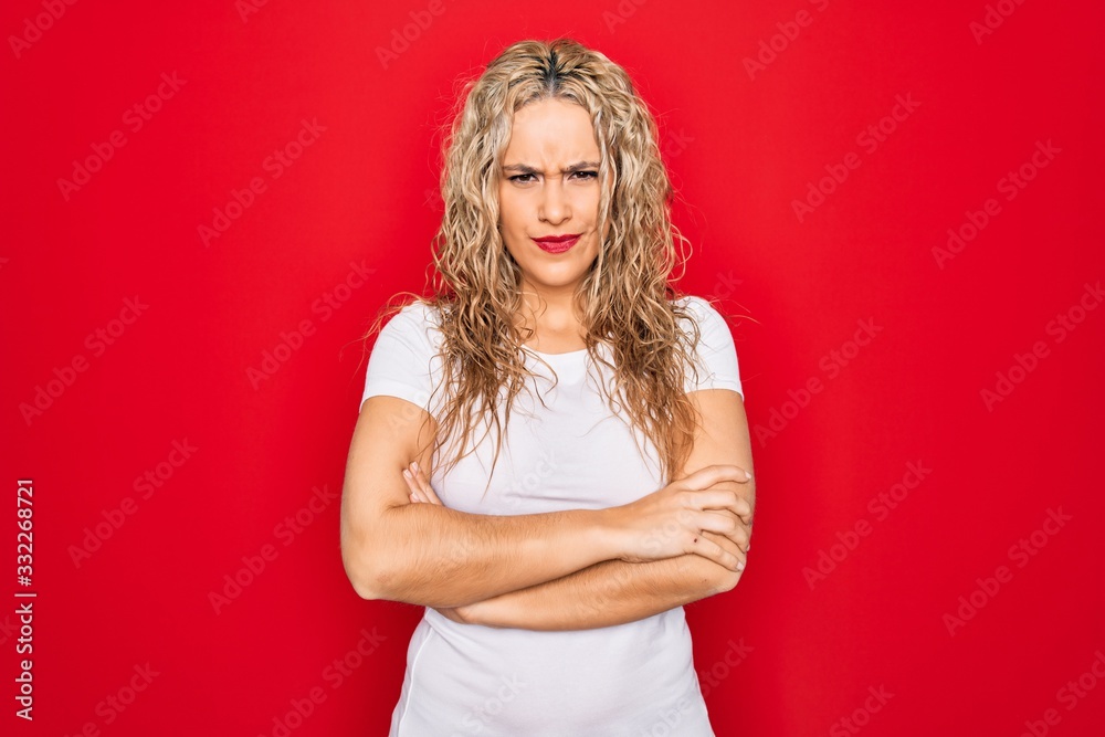 Young beautiful blonde woman wearing casual t-shirt standing over isolated red background skeptic and nervous, disapproving expression on face with crossed arms. Negative person. Young beautiful blonde woman wearing casual t-shirt standing over isolated red background skeptic and nervous, disapproving expression on face with crossed arms. Negative person.