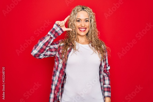 Young beautiful blonde woman wearing casual shirt standing over isolated red background smiling and confident gesturing with hand doing small size sign with fingers looking and the camera. Measure