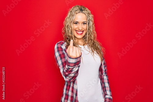 Young beautiful blonde woman wearing casual shirt standing over isolated red background Beckoning come here gesture with hand inviting welcoming happy and smiling