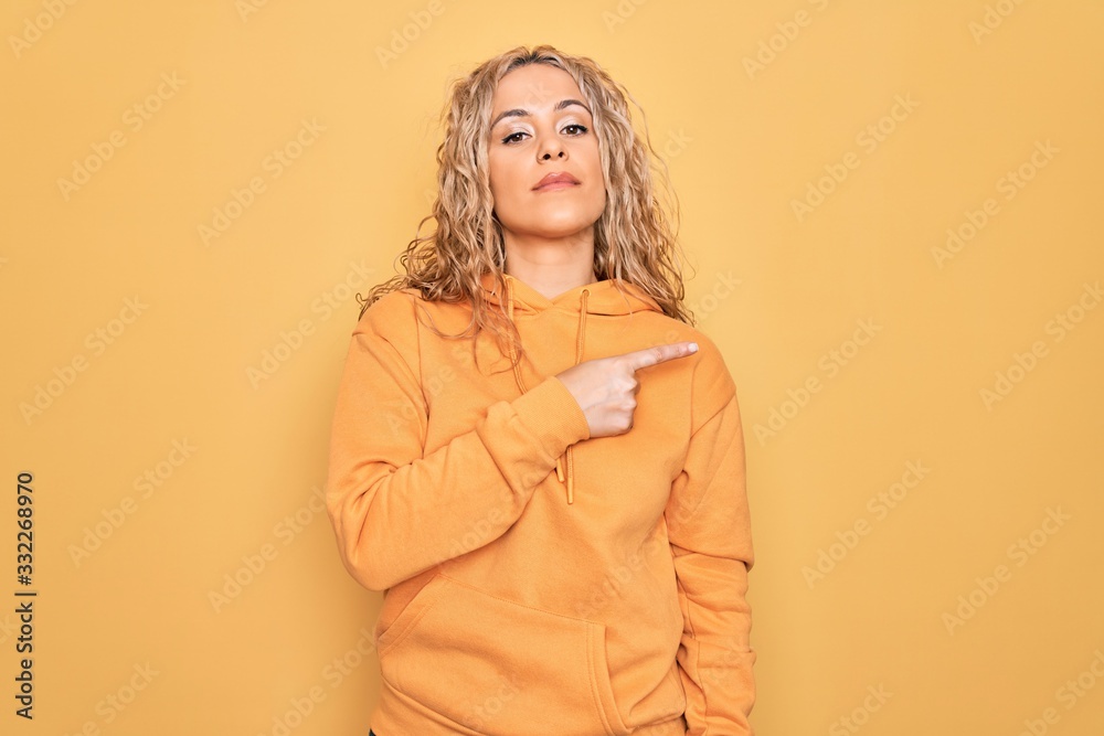 Young beautiful blonde sporty woman wearing casual sweatshirt over yellow background Pointing with hand finger to the side showing advertisement, serious and calm face Young beautiful blonde sporty woman wearing casual sweatshirt over yellow background Pointing with hand finger to the side showing advertisement, serious and calm face