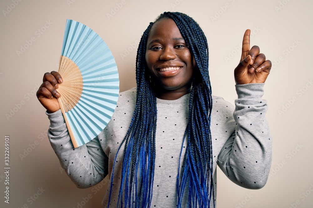 Young african american plus size woman with braids using hand fan over white background surprised with an idea or question pointing finger with happy face, number one Young african american plus size woman with braids using hand fan over white background surprised with an idea or question pointing finger with happy face, number one