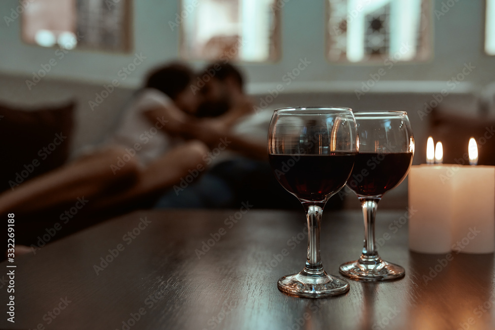 Romance never gets old. Wine glasses and candles on table with couple relaxing in the background on couch