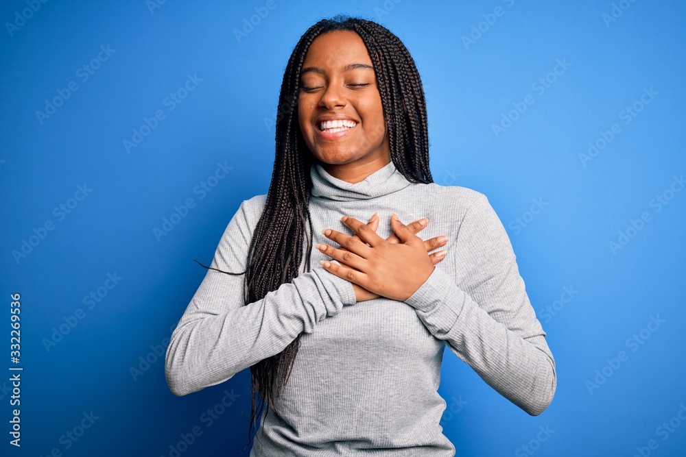 Young african american woman standing wearing casual turtleneck over blue isolated background smiling with hands on chest with closed eyes and grateful gesture on face. Health concept. Young african american woman standing wearing casual turtleneck over blue isolated background smiling with hands on chest with closed eyes and grateful gesture on face. Health concept.