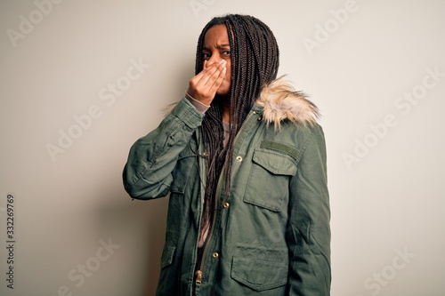 Young african american woman wearing winter parka coat over isolated background smelling something stinky and disgusting, intolerable smell, holding breath with fingers on nose. Bad smell