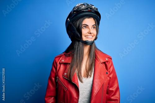 Young beautiful brunette motorcycliste woman wearing motorcycle helmet and jacket looking away to side with smile on face, natural expression. Laughing confident. © Krakenimages.com