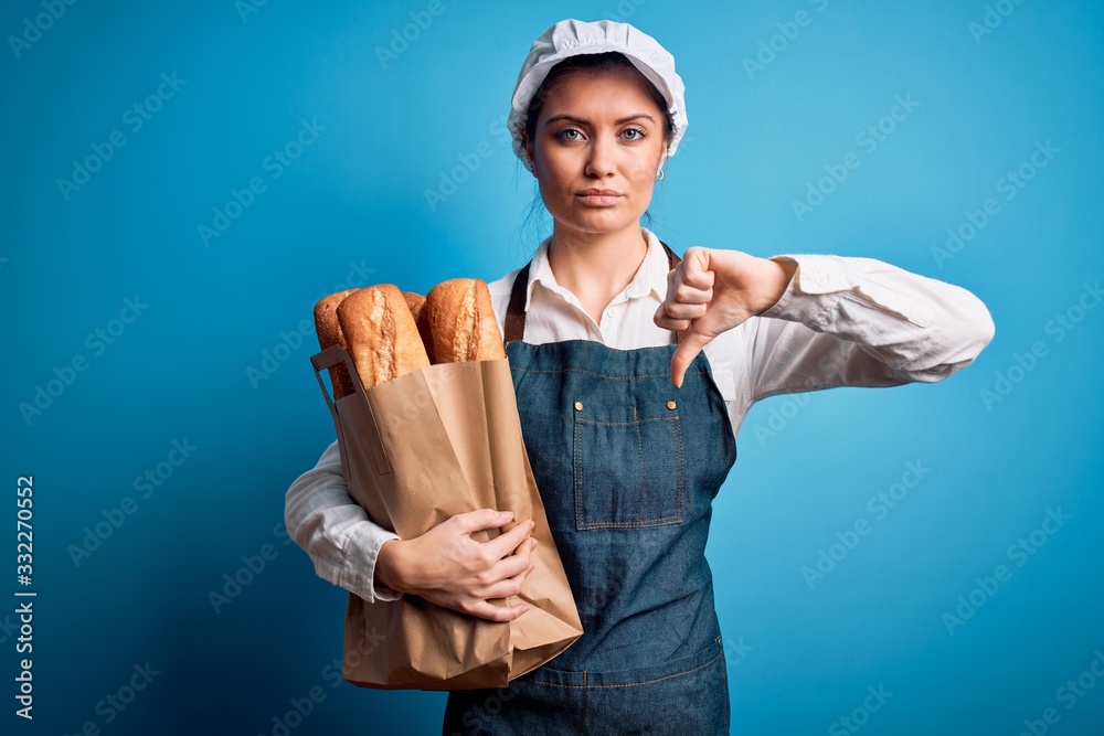 Young beautiful baker woman with blue eyes holding paper bag with  bread with angry face, negative sign showing dislike with thumbs down, rejection concept