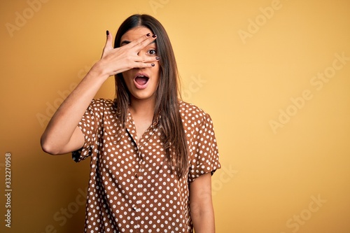 Young beautiful brunette woman wearing casual shirt over isolated yellow background peeking in shock covering face and eyes with hand  looking through fingers with embarrassed expression.