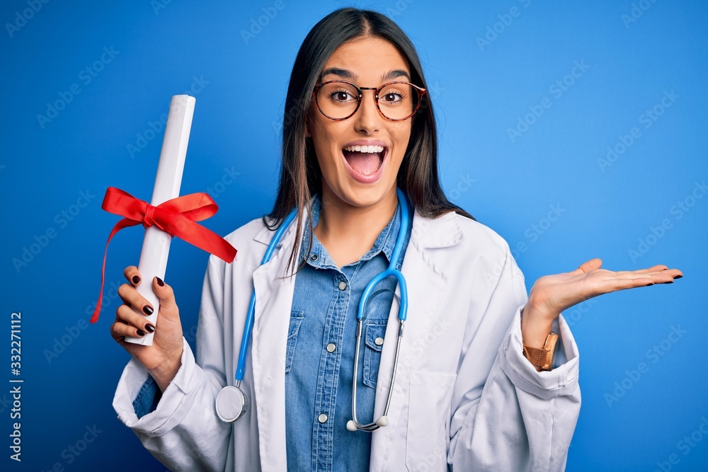 Young beautiful brunette doctor woman wearing glasses and coat holding diploma degree very happy and excited, winner expression celebrating victory screaming with big smile and raised hands