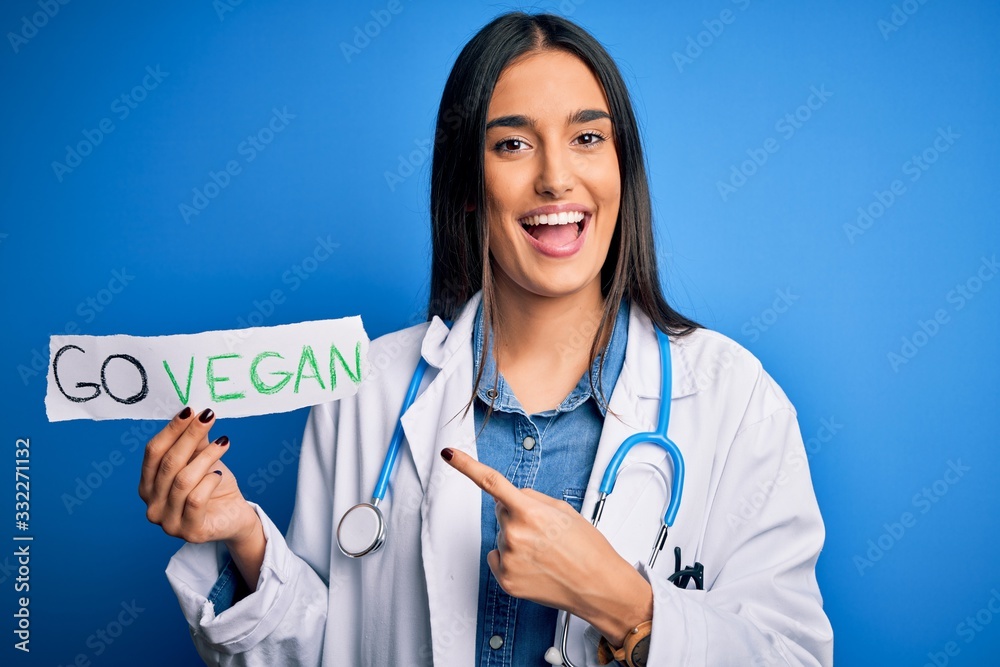 Young beautiful brunette doctor woman wearing coat holding paper with go vegan message very happy pointing with hand and finger Young beautiful brunette doctor woman wearing coat holding paper with go vegan message very happy pointing with hand and finger