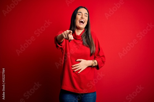 Young beautiful brunette woman wearing casual sweater over isolated red background laughing at you, pointing finger to the camera with hand over body, shame expression