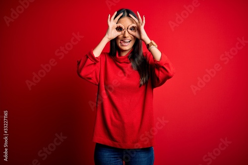 Young beautiful brunette woman wearing casual sweater over isolated red background doing ok gesture like binoculars sticking tongue out, eyes looking through fingers. Crazy expression.