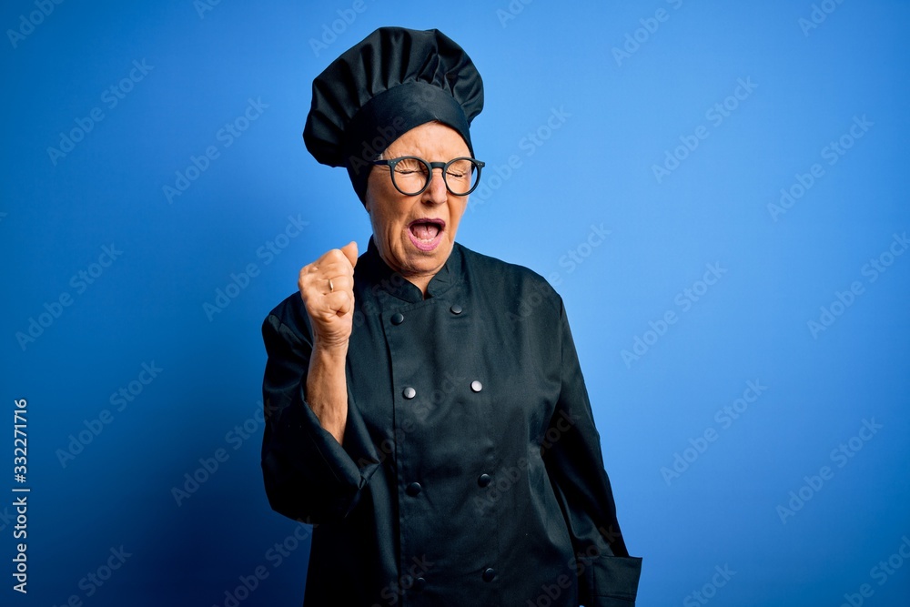 Senior beautiful grey-haired chef woman wearing cooker uniform and hat over blue background angry and mad raising fist frustrated and furious while shouting with anger. Rage and aggressive concept. Senior beautiful grey-haired chef woman wearing cooker uniform and hat over blue background angry and mad raising fist frustrated and furious while shouting with anger. Rage and aggressive concept.