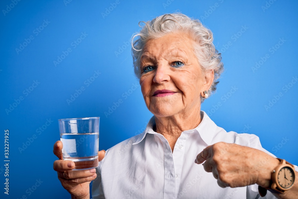 Senior beautiful woman drinking glass of water standing over isolated blue background with surprise face pointing finger to himself Senior beautiful woman drinking glass of water standing over isolated blue background with surprise face pointing finger to himself