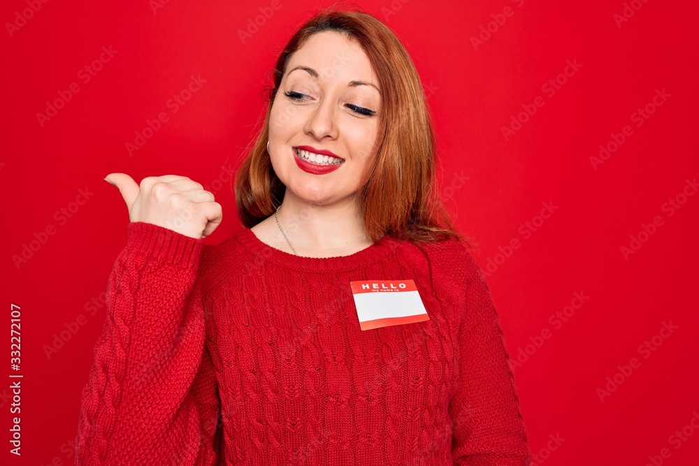 Young beautiful redhead woman wearing sticker presentation with hello my name message smiling with happy face looking and pointing to the side with thumb up.