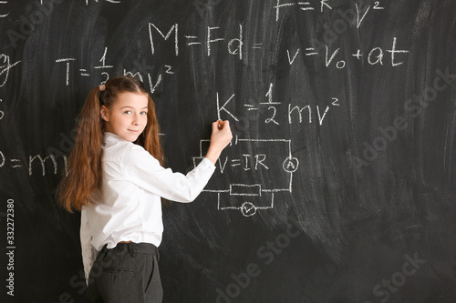 Cute little girl writing on blackboard at physics lesson in classroom