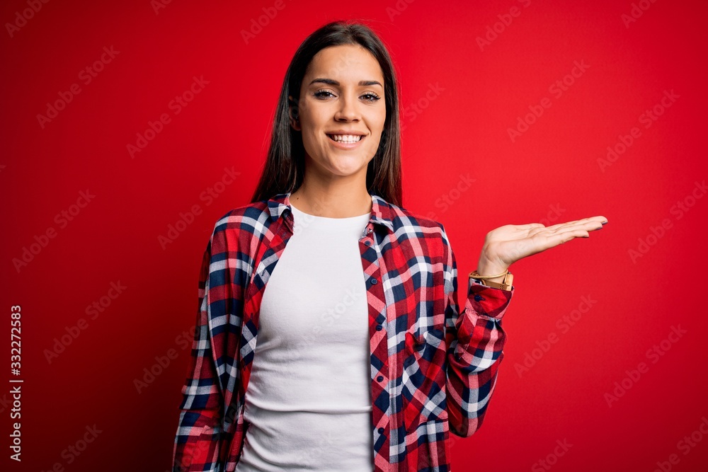 Young beautiful brunette woman wearing casual shirt standing over isolated red background smiling cheerful presenting and pointing with palm of hand looking at the camera. Young beautiful brunette woman wearing casual shirt standing over isolated red background smiling cheerful presenting and pointing with palm of hand looking at the camera.