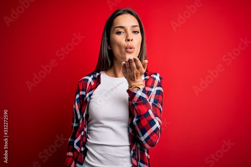 Young beautiful brunette woman wearing casual shirt standing over isolated red background looking at the camera blowing a kiss with hand on air being lovely and sexy. Love expression.