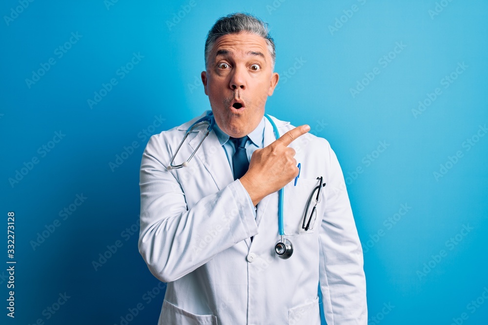 Middle age handsome grey-haired doctor man wearing coat and blue stethoscope Surprised pointing with finger to the side, open mouth amazed expression. Middle age handsome grey-haired doctor man wearing coat and blue stethoscope Surprised pointing with finger to the side, open mouth amazed expression.
